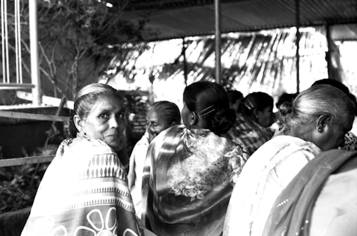 A group of women, primarily older, are sitting closely together in an outdoor setting with a thatched roof above them. They are wearing traditional attire with intricate patterns and appear to be engaged in an event or gathering. One woman in the foreground is facing the camera with a calm expression, while others are turned away or engaged with one another.