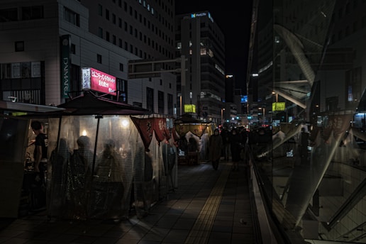 A bustling urban street at night with people gathered in a dimly lit outdoor restaurant or food stall covered with transparent sheets. Tall buildings, including one with a visible MetLife sign, surround the area. Reflections in the glass accentuate the lively yet intimate scene.