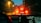 The view through a car windshield on a rainy evening, with focus on droplets of rain. Red brake lights of a car in front brighten the scene, surrounded by dim urban lighting. A navigation system is visible on the dashboard, showing a digital map.