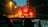 The view through a car windshield on a rainy evening, with focus on droplets of rain. Red brake lights of a car in front brighten the scene, surrounded by dim urban lighting. A navigation system is visible on the dashboard, showing a digital map.