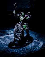 A detailed sculpture of a fantastical warrior figure is posed holding a weapon aloft. The figure appears to be armored, with a dark color scheme accented by green highlights. It stands on a textured, possibly organic, base that looks like a cross between rock and scales. The background is dark, suggesting an ominous or dramatic setting.