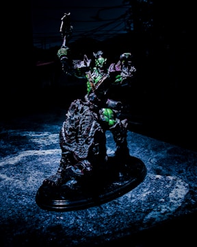 A detailed sculpture of a fantastical warrior figure is posed holding a weapon aloft. The figure appears to be armored, with a dark color scheme accented by green highlights. It stands on a textured, possibly organic, base that looks like a cross between rock and scales. The background is dark, suggesting an ominous or dramatic setting.