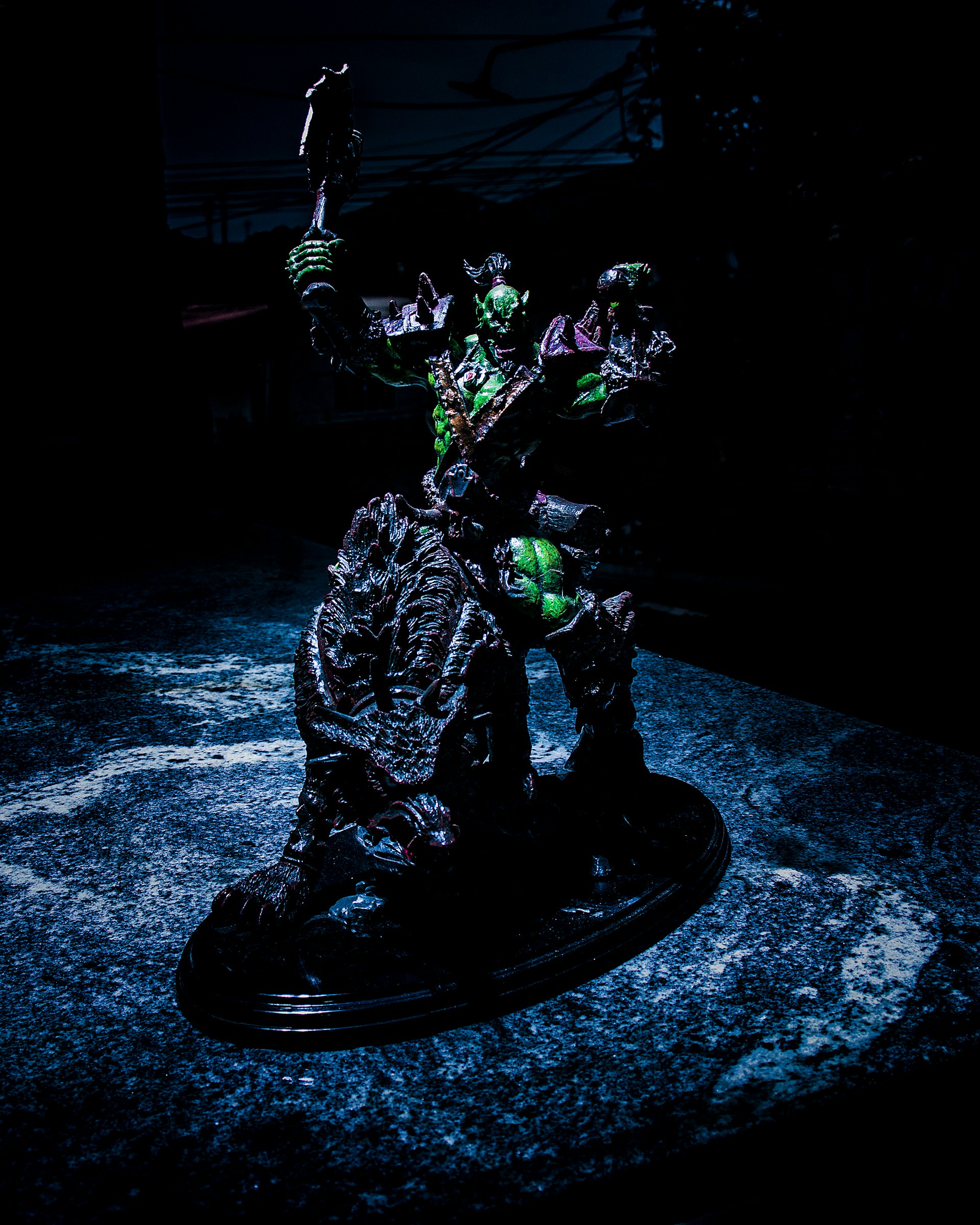 A detailed sculpture of a fantastical warrior figure is posed holding a weapon aloft. The figure appears to be armored, with a dark color scheme accented by green highlights. It stands on a textured, possibly organic, base that looks like a cross between rock and scales. The background is dark, suggesting an ominous or dramatic setting.
