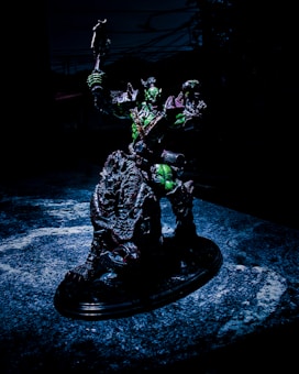 A detailed sculpture of a fantastical warrior figure is posed holding a weapon aloft. The figure appears to be armored, with a dark color scheme accented by green highlights. It stands on a textured, possibly organic, base that looks like a cross between rock and scales. The background is dark, suggesting an ominous or dramatic setting.