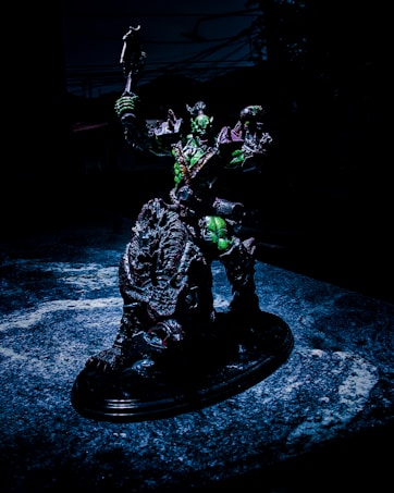 A detailed sculpture of a fantastical warrior figure is posed holding a weapon aloft. The figure appears to be armored, with a dark color scheme accented by green highlights. It stands on a textured, possibly organic, base that looks like a cross between rock and scales. The background is dark, suggesting an ominous or dramatic setting.