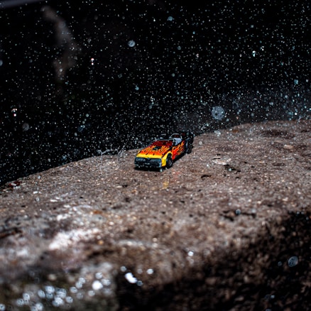 A small toy car with flame decals is positioned on a wet and rough stone surface. Raindrops are visible, creating scattered droplets against a dark background.