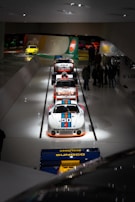 A lineup of classic race cars is displayed in a museum setting, with a focus on a white Porsche adorned with Martini Racing livery and the number 50. The cars are positioned on a sleek, glossy white floor with overhead lighting. Several people are observing the cars in the background, while another brightly colored car is visible near a wall with automotive branding and logos.