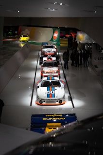 A lineup of classic race cars is displayed in a museum setting, with a focus on a white Porsche adorned with Martini Racing livery and the number 50. The cars are positioned on a sleek, glossy white floor with overhead lighting. Several people are observing the cars in the background, while another brightly colored car is visible near a wall with automotive branding and logos.
