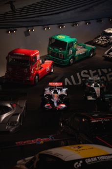 A collection of racing vehicles, including two large trucks in red and green and several race cars, are displayed in an indoor automotive exhibit. The vehicles are arranged in a dimly lit setting, highlighting their designs and logos.