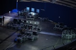 A large number of black equipment cases are stacked and organized in a dimly lit backstage area of an auditorium or stadium. The space is mostly empty, with some seating visible in the upper right corner. An individual is seen on the left side, possibly working with equipment.