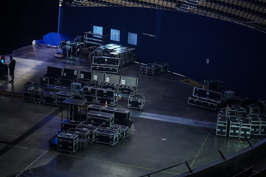A large number of black equipment cases are stacked and organized in a dimly lit backstage area of an auditorium or stadium. The space is mostly empty, with some seating visible in the upper right corner. An individual is seen on the left side, possibly working with equipment.