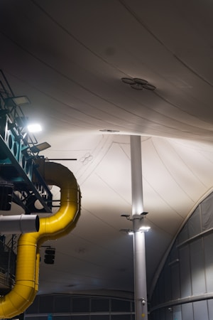 A large, industrial indoor setting featuring a prominent yellow ventilation duct extending across the ceiling. The area is lit by overhead lights attached to tall, white support beams. The ceiling consists of large, white, fabric-like panels and there are some additional metallic structures present.