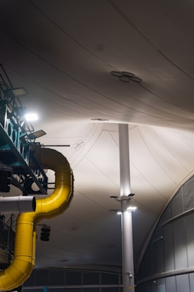 A large, industrial indoor setting featuring a prominent yellow ventilation duct extending across the ceiling. The area is lit by overhead lights attached to tall, white support beams. The ceiling consists of large, white, fabric-like panels and there are some additional metallic structures present.