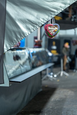 A small heart-shaped gingerbread with colorful icing hangs prominently in the foreground. The words 'Jel&ouml;t&uuml;d, biz&rsquo; saf' are written on it. Behind, a blurred setting of a food stall can be seen, with some people in the background and lights that suggest an outdoor market or festive environment.