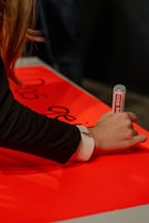 A person is writing on a bright red surface using a silver and white marker. The individual's hand and part of the arm are visible, wearing a black sleeve with a white cuff. Some black text is partially visible on the red surface.