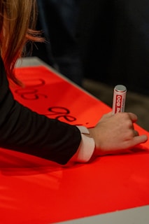 A person is writing on a bright red surface using a silver and white marker. The individual's hand and part of the arm are visible, wearing a black sleeve with a white cuff. Some black text is partially visible on the red surface.