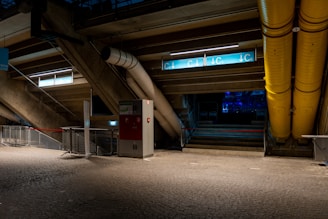 A dimly lit interior space, possibly part of a stadium or industrial area, featuring concrete structures, metal railings, and a mix of pipes. The ground is paved with textured cobblestone-like tiles. There are signs with blue and white direction markers indicating C3, C4, and C5. The image also includes large yellow ducts and a red and white metal cabinet.