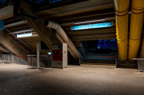 A dimly lit interior space, possibly part of a stadium or industrial area, featuring concrete structures, metal railings, and a mix of pipes. The ground is paved with textured cobblestone-like tiles. There are signs with blue and white direction markers indicating C3, C4, and C5. The image also includes large yellow ducts and a red and white metal cabinet.