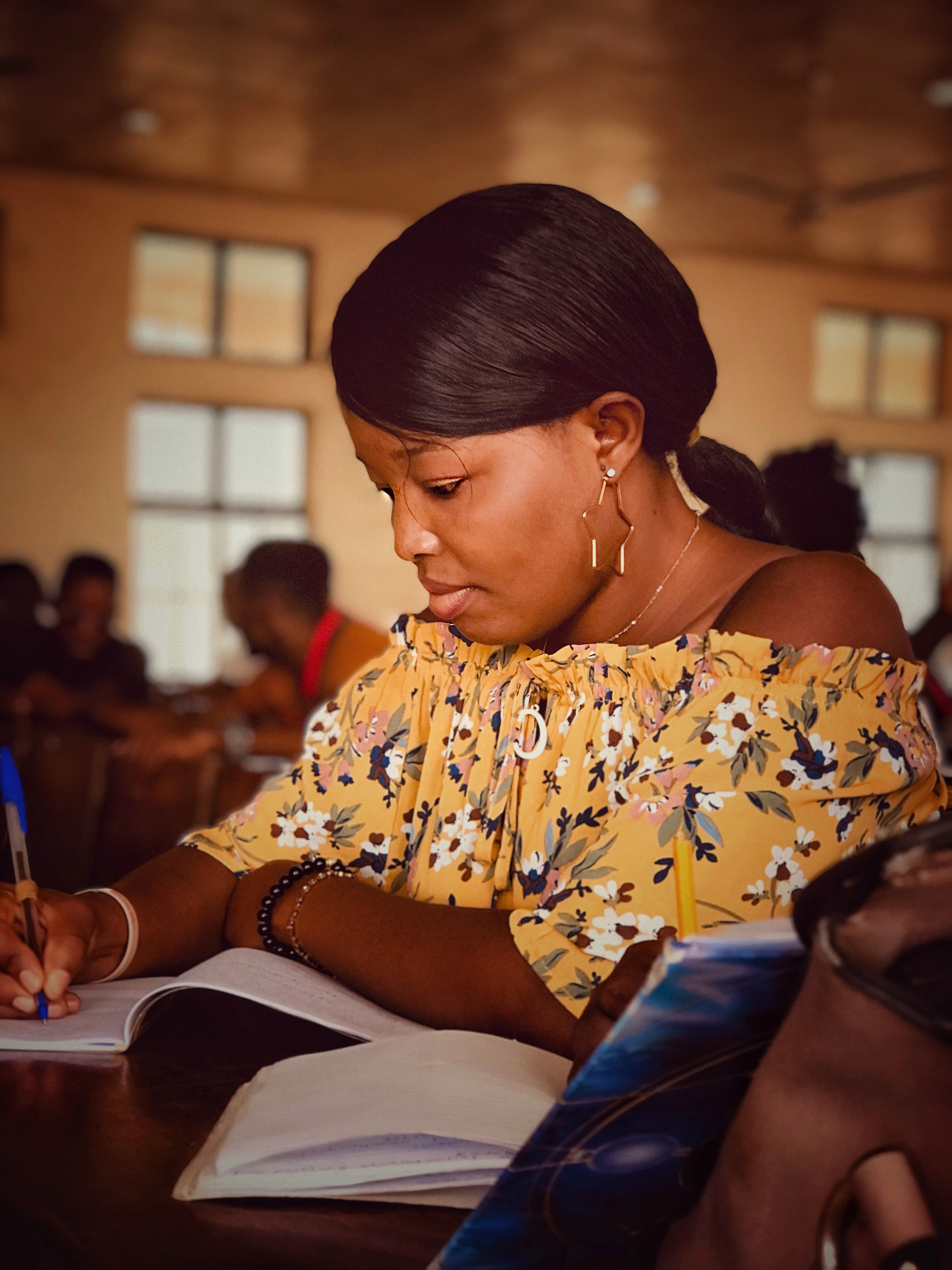 A woman in a yellow floral top writing in an exercise book at a desk
