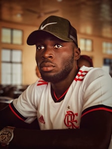 A person is wearing a white and red sports jersey with logos and is seated indoors. They have a short beard and are wearing a dark green cap. The background features a softly lit room with blurred windows and ceiling fans.