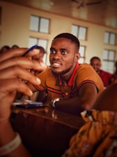 A man in a brown polo shirt with black and red stripes is smiling and leaning forward slightly, appearing engaged. He holds a pen, possibly participating in a conversation. Several blurred figures are in the background, seated in a room with large windows.