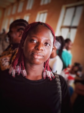 A young woman with red braided hair and a black shirt is standing indoors, looking directly at the camera. The background is a bit blurred, with a few other people and some windows visible.