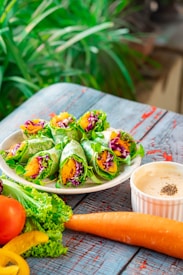 A plate of fresh vegetable wraps filled with greens, shredded purple cabbage, and julienned carrots is set on a rustic wooden table. Alongside the plate, there is a white ramekin filled with creamy dipping sauce sprinkled with black pepper. Accompanying the wraps are additional fresh ingredients including a whole carrot, a ripe tomato, yellow bell pepper slices, and lettuce leaves. The background is lush with green foliage, adding a vibrant and natural setting to the scene.