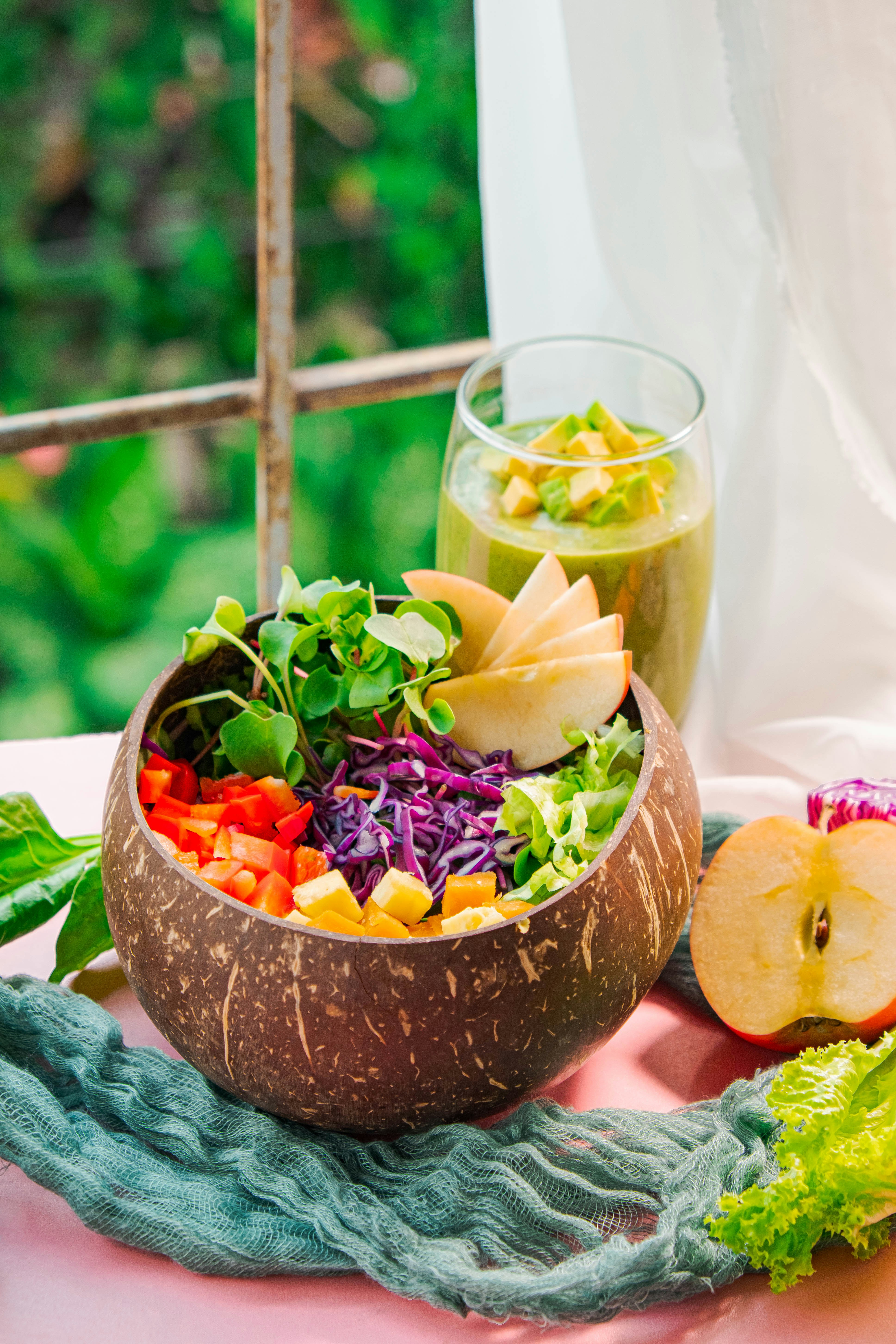 A wooden bowl is filled with a colorful assortment of fresh vegetables and fruits, including red peppers, shredded purple cabbage, wedge-shaped apple slices, and leafy greens. Beside the bowl, a glass of green smoothie is partially filled and garnished with small fruit cubes. The setting appears to be a casual meal likely influenced by nature, as a slice of apple and leafy greens are placed around the bowl on a textured green fabric, with a view of lush greenery visible through a window.