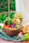 A wooden bowl is filled with a colorful assortment of fresh vegetables and fruits, including red peppers, shredded purple cabbage, wedge-shaped apple slices, and leafy greens. Beside the bowl, a glass of green smoothie is partially filled and garnished with small fruit cubes. The setting appears to be a casual meal likely influenced by nature, as a slice of apple and leafy greens are placed around the bowl on a textured green fabric, with a view of lush greenery visible through a window.