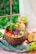 A wooden bowl is filled with a colorful assortment of fresh vegetables and fruits, including red peppers, shredded purple cabbage, wedge-shaped apple slices, and leafy greens. Beside the bowl, a glass of green smoothie is partially filled and garnished with small fruit cubes. The setting appears to be a casual meal likely influenced by nature, as a slice of apple and leafy greens are placed around the bowl on a textured green fabric, with a view of lush greenery visible through a window.