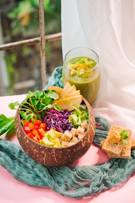 A wooden bowl filled with a colorful assortment of fresh vegetables, including diced red peppers, sliced avocado, shredded purple cabbage, and microgreens, accompanied by cubed bread pieces. Beside the bowl is a glass of green smoothie garnished with diced avocado, placed on a textured blue fabric atop a pink surface. Slices of apple are arranged in the bowl, and there are two pieces of whole-grain bread with a spread and microgreens nearby.