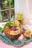 A wooden bowl filled with a colorful assortment of fresh vegetables, including diced red peppers, sliced avocado, shredded purple cabbage, and microgreens, accompanied by cubed bread pieces. Beside the bowl is a glass of green smoothie garnished with diced avocado, placed on a textured blue fabric atop a pink surface. Slices of apple are arranged in the bowl, and there are two pieces of whole-grain bread with a spread and microgreens nearby.