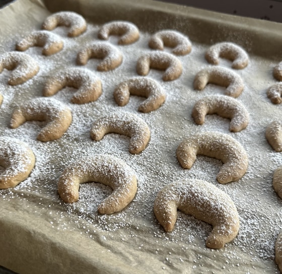 A baking tray is filled with crescent-shaped cookies evenly dusted with powdered sugar. The treats are arranged in neat rows on brown parchment paper.