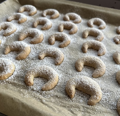 A baking tray is filled with crescent-shaped cookies evenly dusted with powdered sugar. The treats are arranged in neat rows on brown parchment paper.