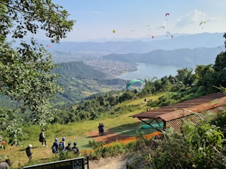 Paragliders fill the sky above a scenic viewpoint overlooking a vast expanse of lush green hills and a large lake bordered by a city. People with helmets gather at a launching area, surrounded by greenery and trees.
