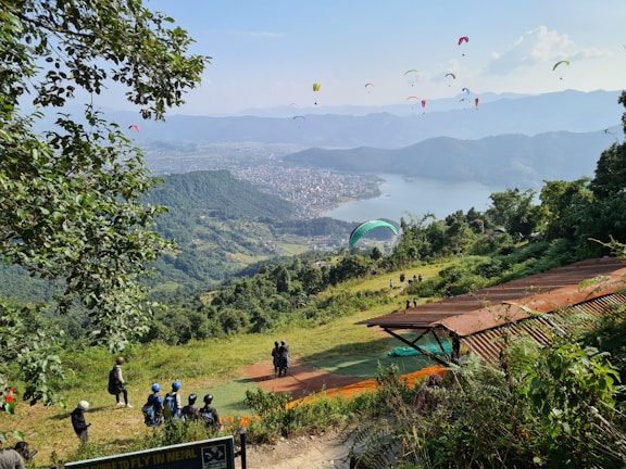 Paragliders fill the sky above a scenic viewpoint overlooking a vast expanse of lush green hills and a large lake bordered by a city. People with helmets gather at a launching area, surrounded by greenery and trees.