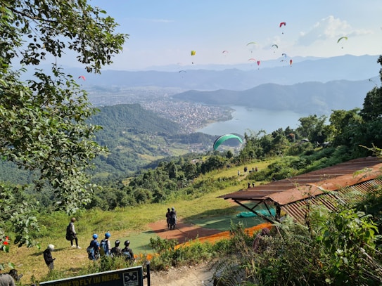 Paragliders fill the sky above a scenic viewpoint overlooking a vast expanse of lush green hills and a large lake bordered by a city. People with helmets gather at a launching area, surrounded by greenery and trees.