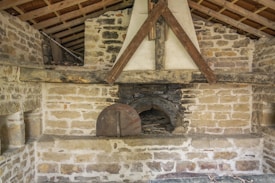 A rustic brick wall structure featuring an old-fashioned stone and clay fireplace or oven with a wooden beam support. The structure is set in a sloped-roof interior, with visible timber beams supporting the roof.