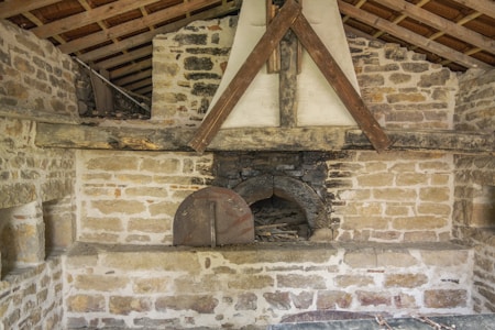 A rustic brick wall structure featuring an old-fashioned stone and clay fireplace or oven with a wooden beam support. The structure is set in a sloped-roof interior, with visible timber beams supporting the roof.