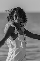 A person with curly hair and sunglasses is smiling and wearing a sleeveless dress with ruffled details, standing outdoors near a body of water. The image is black and white, capturing a casual, candid moment.