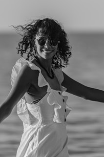 A person with curly hair and sunglasses is smiling and wearing a sleeveless dress with ruffled details, standing outdoors near a body of water. The image is black and white, capturing a casual, candid moment.