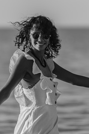 A person with curly hair and sunglasses is smiling and wearing a sleeveless dress with ruffled details, standing outdoors near a body of water. The image is black and white, capturing a casual, candid moment.