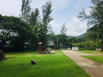 A serene park setting with vibrant green grass and tall trees surrounding a small wooden play structure. There is a pathway leading towards a white building in the background, bordered by a metal fence. A flagpole stands near the building, and there are hills visible in the distance.