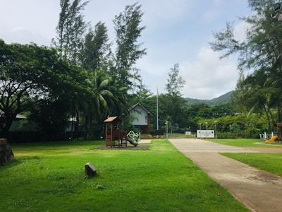 A serene park setting with vibrant green grass and tall trees surrounding a small wooden play structure. There is a pathway leading towards a white building in the background, bordered by a metal fence. A flagpole stands near the building, and there are hills visible in the distance.