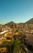 A sunny landscape featuring a complex of white, modern buildings set against a backdrop of rugged hills. The architecture is surrounded by palm trees and desert shrubs, reflecting a desert resort environment. The sky is clear and blue, contributing to a serene and warm atmosphere.