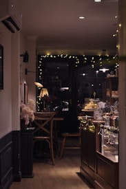 A cozy café interior with dim lighting and warm ambiance. There's a wooden table and chairs set near a window adorned with string lights. A counter to the right displays pastries under glass cloches, surrounded by various cutlery and condiments. The space is decorated with flowers and soft lighting from a table lamp.