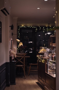 A cozy caf&eacute; interior with dim lighting and warm ambiance. There's a wooden table and chairs set near a window adorned with string lights. A counter to the right displays pastries under glass cloches, surrounded by various cutlery and condiments. The space is decorated with flowers and soft lighting from a table lamp.