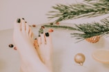 Feet with dark green nail polish are submerged in water, surrounded by festive elements like evergreen branches, a pine cone, small red and brown decorations, and a slice of lemon. A gold ornament floats nearby, creating a serene and holiday-themed atmosphere.