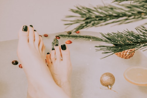 Feet with dark green nail polish are submerged in water, surrounded by festive elements like evergreen branches, a pine cone, small red and brown decorations, and a slice of lemon. A gold ornament floats nearby, creating a serene and holiday-themed atmosphere.