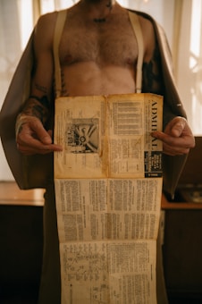 A person with tattoos on their arms and torso is holding an aged and detailed instruction manual or diagram. The paper appears yellowed, suggesting it is old. The person is partially dressed, and the background shows a softly lit indoor setting.